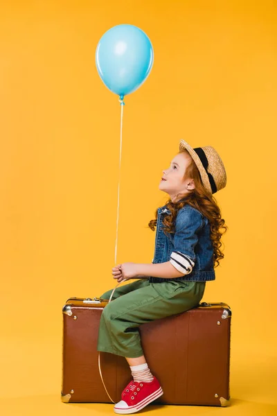 Vista lateral del niño con globo sentado en la maleta aislado en amarillo - foto de stock