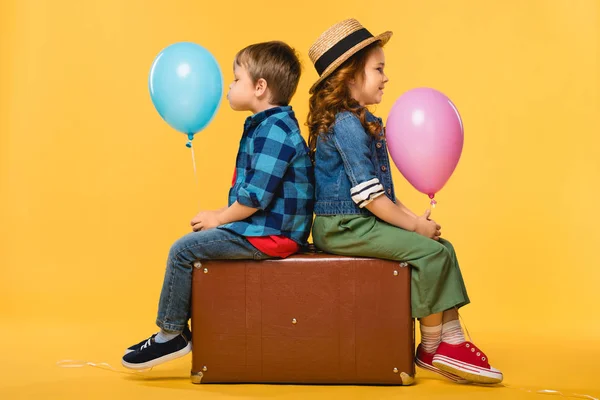 Vista laterale di bambini con palloncini seduti su valigia di pelle isolata su giallo — Foto stock