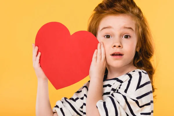 Portrait of shocked child with red paper heart isolated on yellow, st valentines day concept — Stock Photo