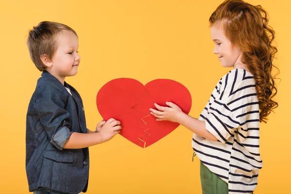 Vista laterale di bambini sorridenti che tengono insieme il cuore di carta rossa isolato sul giallo, concetto di giorno st valentines — Foto stock