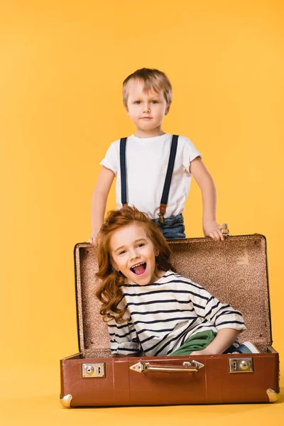 Glückliches Kind im Koffer sitzend mit Junge in der Nähe stehend isoliert auf gelb — Stockfoto