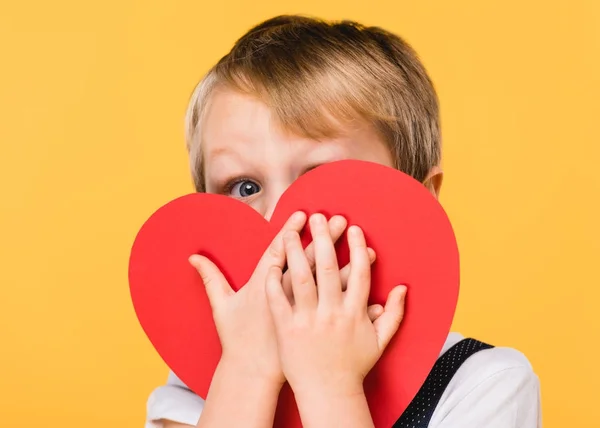 Obscured vista de niño cubriendo la cara con rojo corazón de papel aislado en amarillo, San Valentín concepto de día - foto de stock
