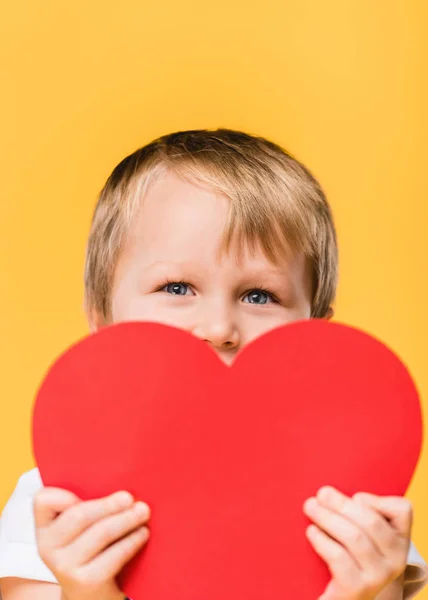 Obscured vista de niño cubriendo la cara con rojo corazón de papel aislado en amarillo, San Valentín concepto de día - foto de stock