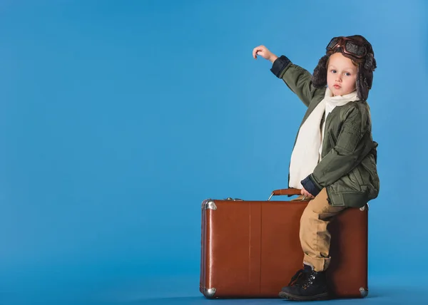 Seitenansicht eines Jungen im Pilotenkostüm, der auf einem Koffer sitzt, isoliert auf blauem Grund — Stockfoto