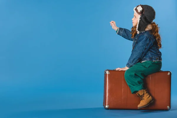 Side view of adorable preteen kid in pilot costume sitting on suitcase isolated on blue — Stock Photo