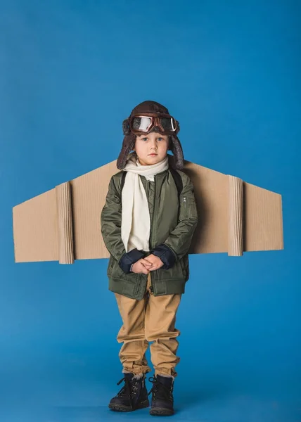 Niedlicher kleiner Junge im Pilotenkostüm mit Papierflugzeugflügel isoliert auf blau — Stockfoto