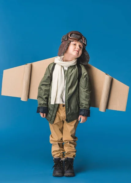 Cute little boy in pilot costume with paper plane wing isolated on blue — Stock Photo