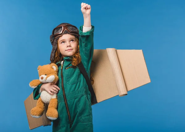Portrait of cute child in pilot costume with teddy bear and handmade paper plane wings isolated on blue — Stock Photo