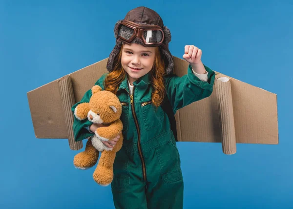 Portrait d'enfant mignon en costume de pilote avec ours en peluche et ailes d'avion en papier à la main isolé sur bleu — Photo de stock