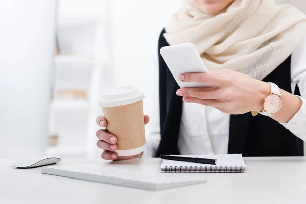 Vista parziale di donna d'affari con caffè per andare utilizzando smartphone sul posto di lavoro — Foto stock