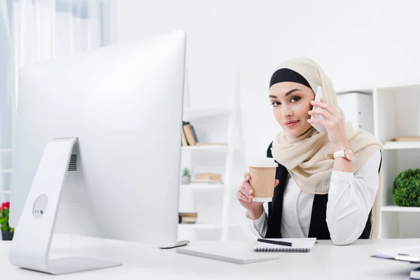 Businesswoman in hijab with coffee to go talking on smartphone at workplace in office — Stock Photo