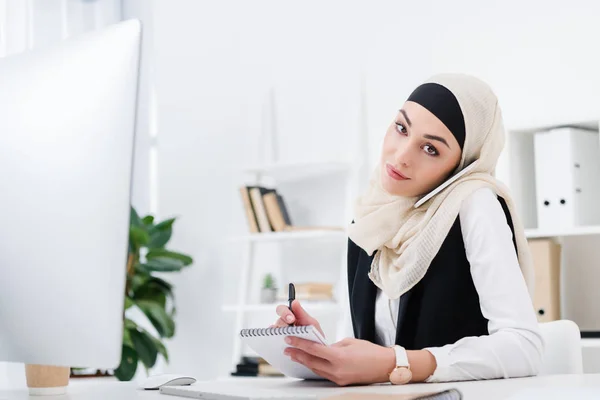 Portrait of arabic businesswoman in hijab with notebook talking on smartphone at workplace in office — Stock Photo