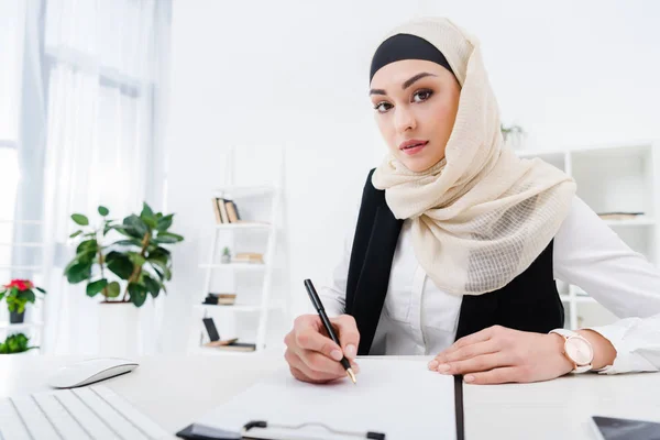 Retrato de mujer de negocios árabe mirando a la cámara mientras firma papeles en el lugar de trabajo - foto de stock