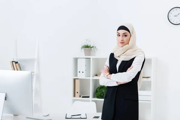 Retrato de mujer de negocios en hiyab con los brazos cruzados de pie en la oficina - foto de stock