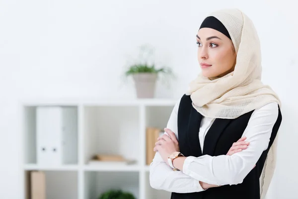 Retrato de mujer de negocios en hiyab con los brazos cruzados de pie en la oficina - foto de stock