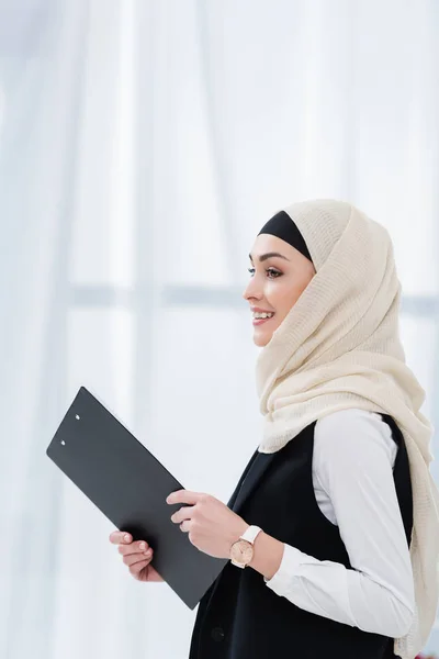 Вид збоку усміхненої арабської бізнес-леді з текою в руках в офісі — стокове фото