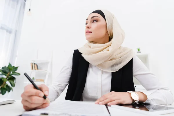 Pensativa mujer de negocios árabe mirando hacia otro lado mientras está sentada en el lugar de trabajo en la oficina - foto de stock