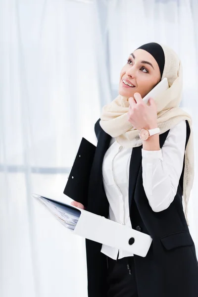 Retrato de mujer de negocios sonriente en hijab con documentos que hablan en el teléfono inteligente en la oficina - foto de stock