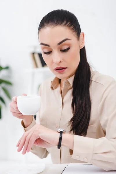 Retrato de mujer de negocios con taza de café tiempo de verificación en el lugar de trabajo en la oficina - foto de stock