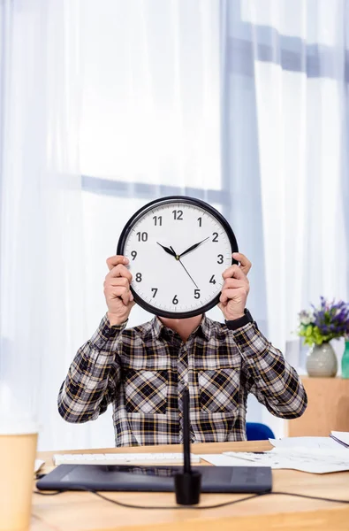 Homme tenant horloge sur le visage tout en étant assis à la table de travail — Photo de stock