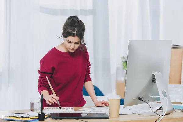 Mujer joven usando paleta de acuarela y cepillo por tabla con computadora - foto de stock