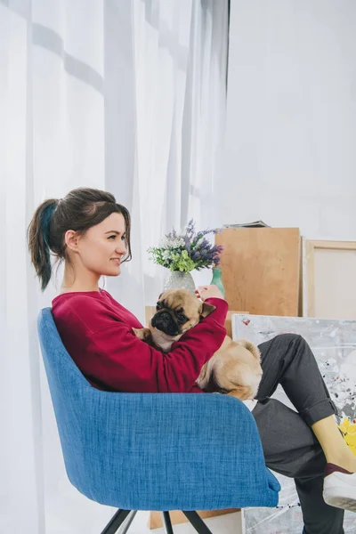 Junges Mädchen kuschelt Mops im Stuhl in modernem Zimmer — Stockfoto