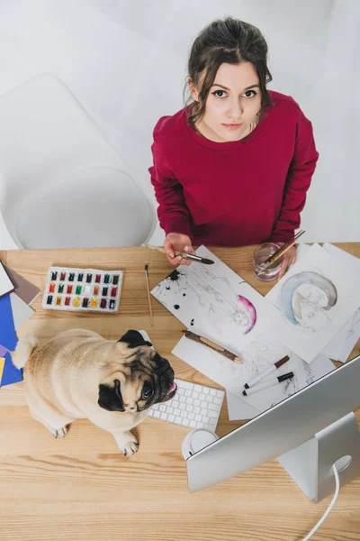 Muchacha joven atractiva trabajando en ilustraciones con lindo pug en la mesa de trabajo con el ordenador - foto de stock