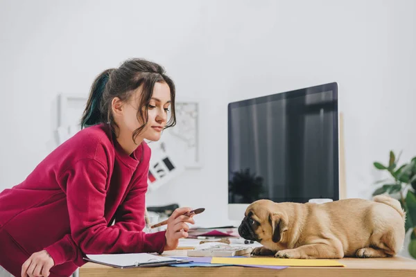 Jovem atraente brincando com pug enquanto trabalhava em ilustrações em home office — Fotografia de Stock