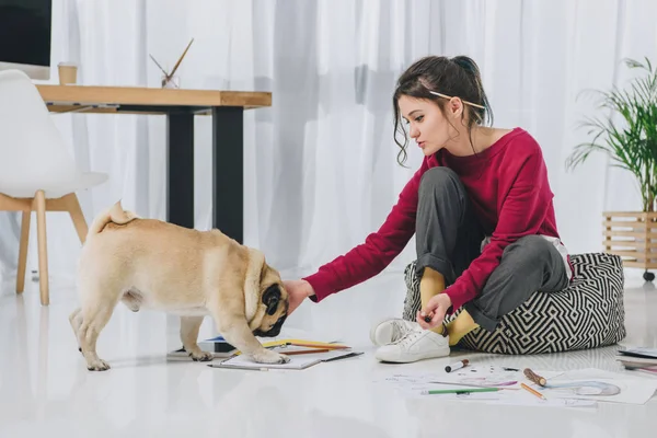 Attractive young girl cuddling pug on floor among sketches — Stock Photo