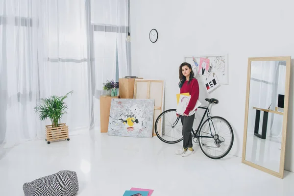 Pretty lady holding sketches and standing by bicycle — Stock Photo