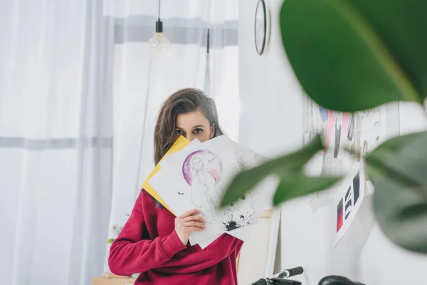 Designer feminina segurando esboços na frente de seu rosto — Fotografia de Stock