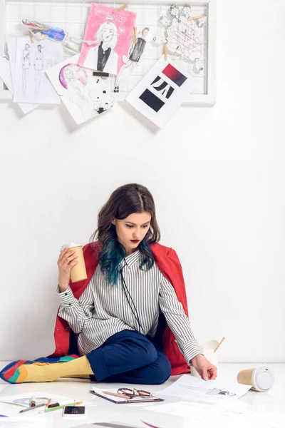 Bonita diseñadora sentada en el suelo con taza de café y trabajando con bocetos - foto de stock