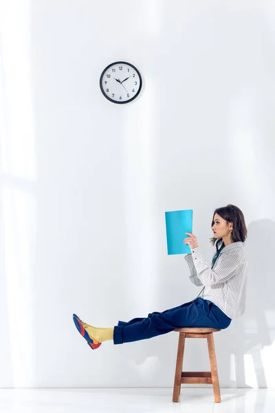 Junge Frau sitzt auf Stuhl und liest Buch unter Uhr — Stockfoto