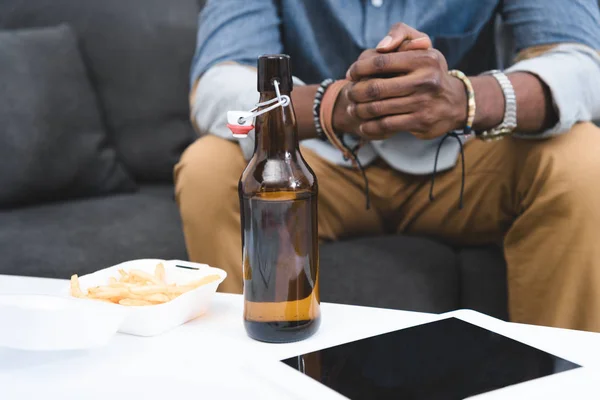 Vista de cerca del chico afroamericano comiendo papas fritas con cerveza en la mesa con tableta digital - foto de stock