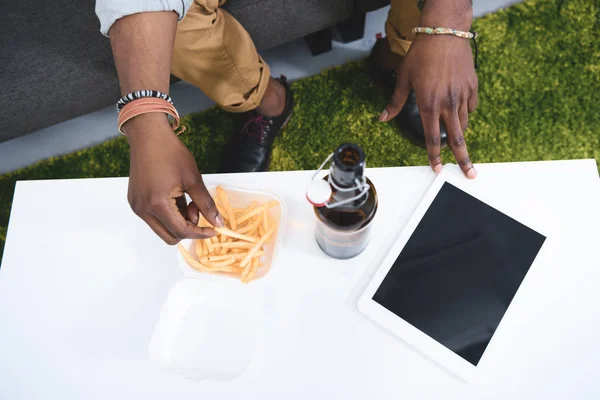 Africam american man eating french fries with beer on table with digital tablet — Stock Photo