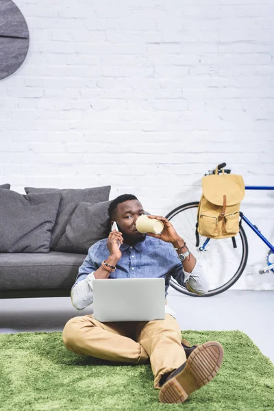 Hombre afroamericano guapo hablando por teléfono y beber café mientras trabaja en el ordenador portátil - foto de stock