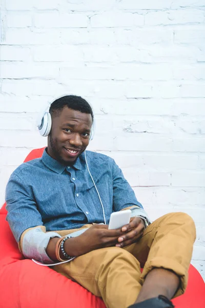 Bel homme afro-américain avec smartphone écoutant de la musique dans des écouteurs et assis dans un sac de haricots — Photo de stock