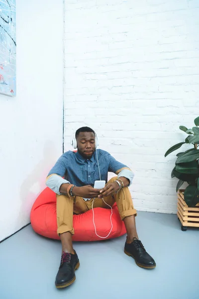 Молодий чоловік зі смартфоном в руках слухає музику в навушниках, сидячи в мішку з бобами — стокове фото