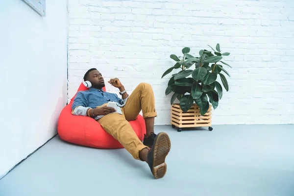 Hombre afroamericano escuchando música en auriculares mientras está sentado en una bolsa de frijoles - foto de stock