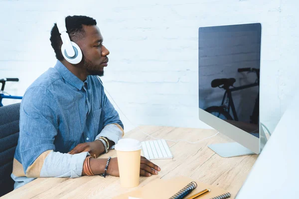 Bonito homem afro-americano em fones de ouvido trabalhando à mesa com computador — Fotografia de Stock