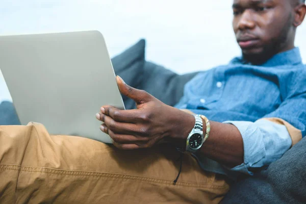 Uomo afroamericano seduto sul divano e al lavoro sul computer portatile — Foto stock