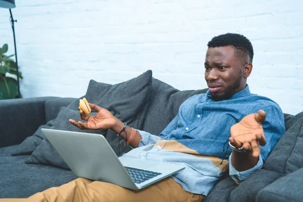 Confundido hombre afroamericano que trabaja en el ordenador portátil mientras está sentado en el sofá y comer hamburguesa - foto de stock