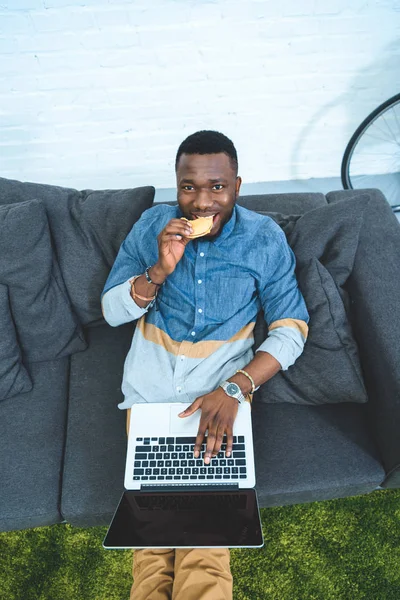 Hombre afroamericano guapo trabajando en el ordenador portátil mientras está sentado en el sofá y comer hamburguesa - foto de stock