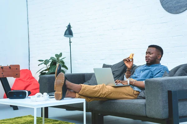 African american man eating hamburger while working on laptop by table with gadgets — Stock Photo