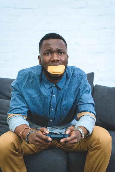 Hombre afroamericano guapo jugando con joystick y sosteniendo hamburguesa en la boca - foto de stock