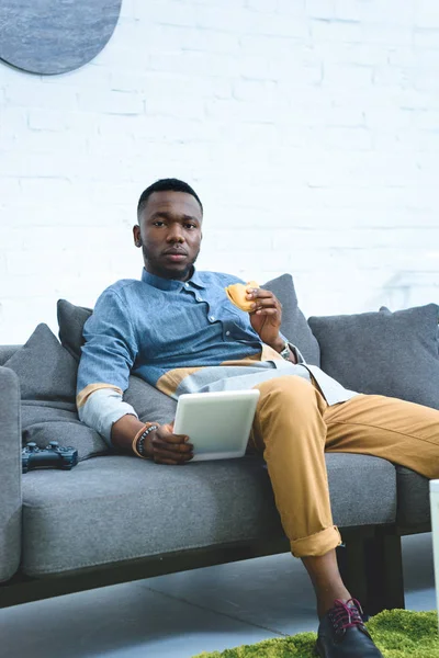 African american man holding tablet and eating hamburger — Stock Photo