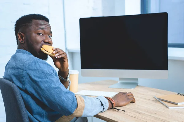 African american man working by computer and eating hamburger — Stock Photo