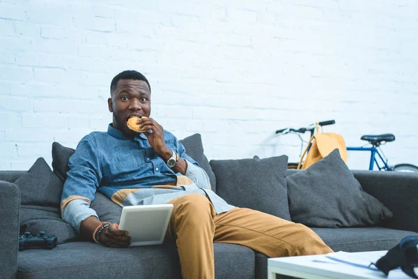 Beau Afro-Américain qui utilise des comprimés et mange du hamburger — Photo de stock