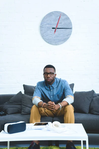 Gadgets modernos en la mesa frente al hombre afroamericano sentado en el sofá - foto de stock