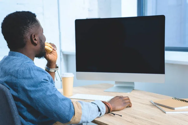 Jovem trabalhando por computador e comendo hambúrguer — Fotografia de Stock